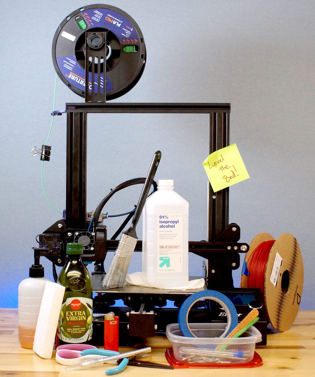 Photo of an Ender 3 Pro surrounded by  olive oil, isopropyl alcohol, dish soap,,  tape, a paint brush and other house hold items